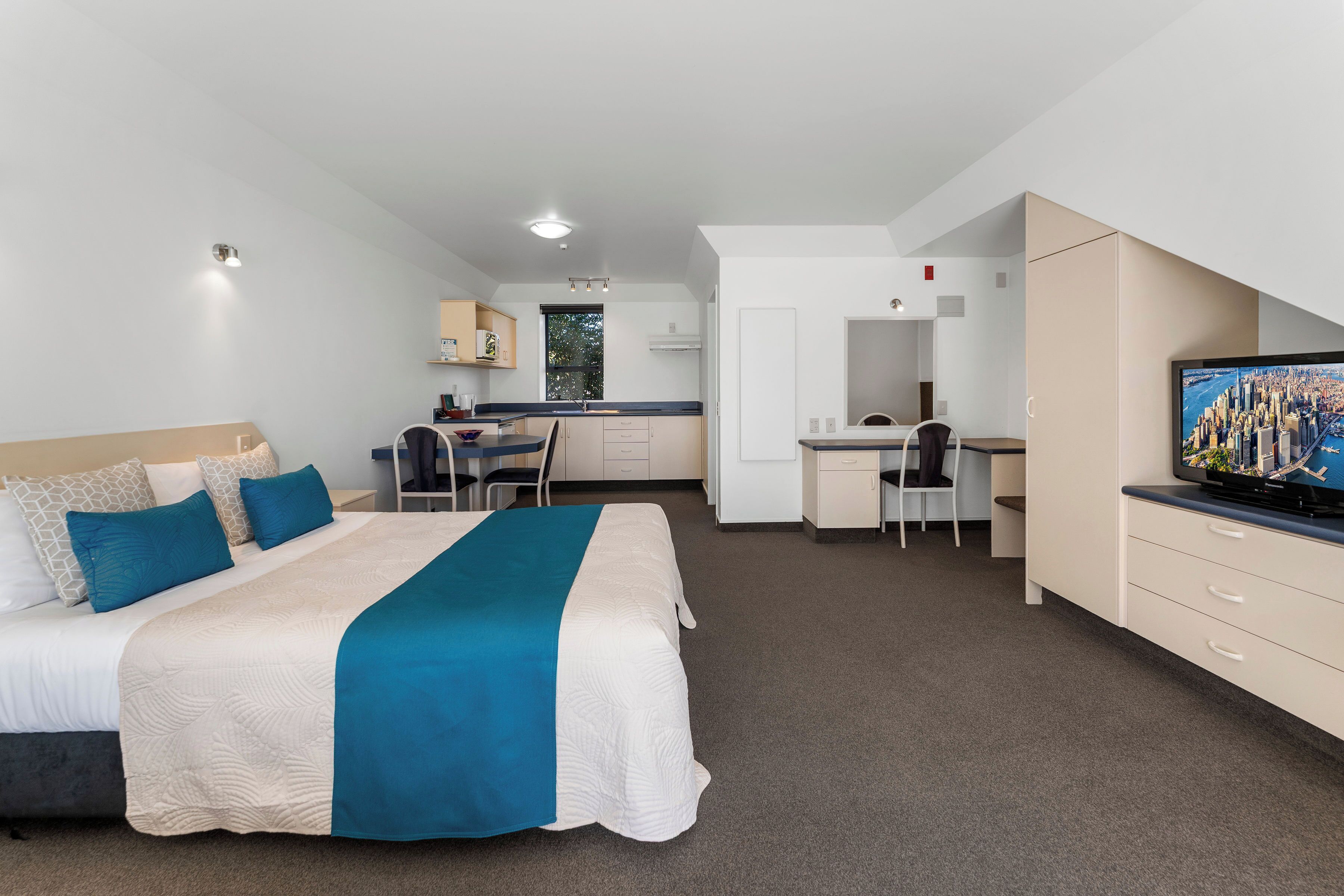 Quarto de hotel moderno com cama, escrivaninha, cozinha compacta e televisão exibindo uma vista urbana.