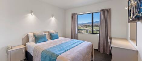 A hotel room with a large bed, a nightstand, a desk, a window with a view of mountains, and a ceiling light.