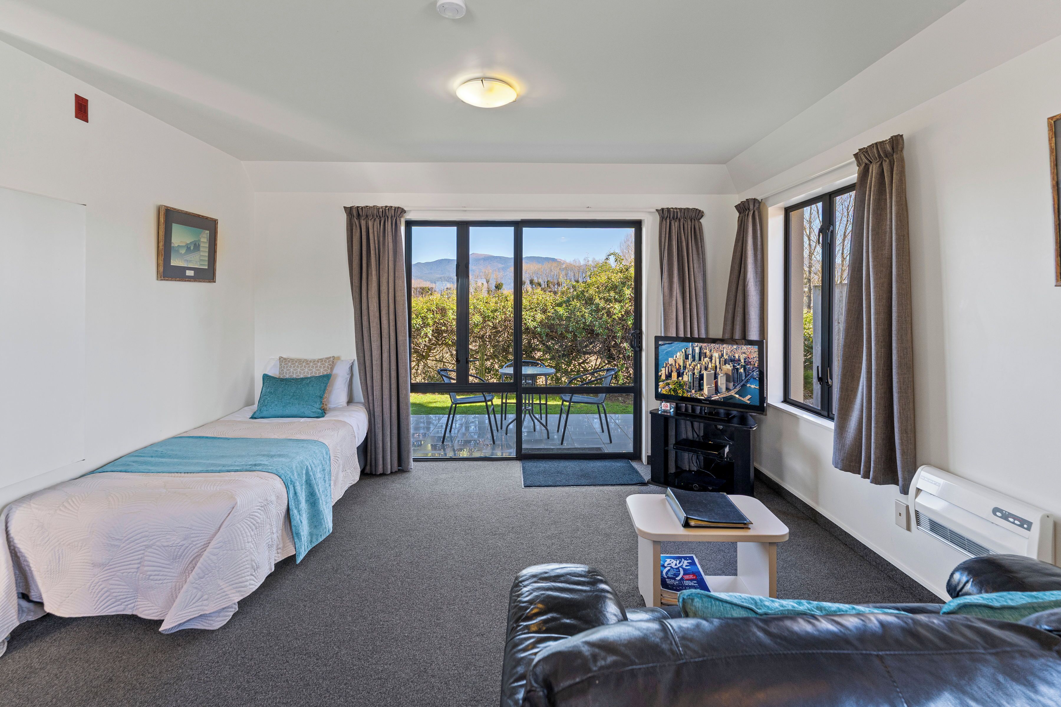 A room with two beds, a sofa, a small table, a TV, and a sliding glass door to an outdoor area with a view of trees and hills.