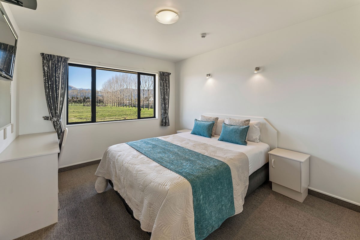 A bedroom with a large bed, a bedside table, a nightstand, a window with curtains, and a view of a grassy field.