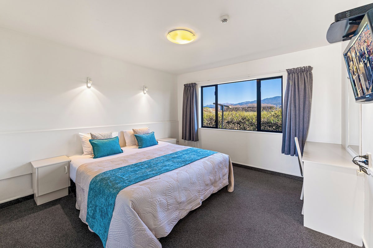 A hotel room with a large bed, a desk, a window with a view of the mountains, and a flat-screen TV mounted on the wall.