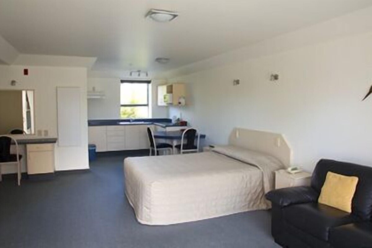 Habitación de hotel compacta con cama, escritorio, silla y una kitchenette pequeña.