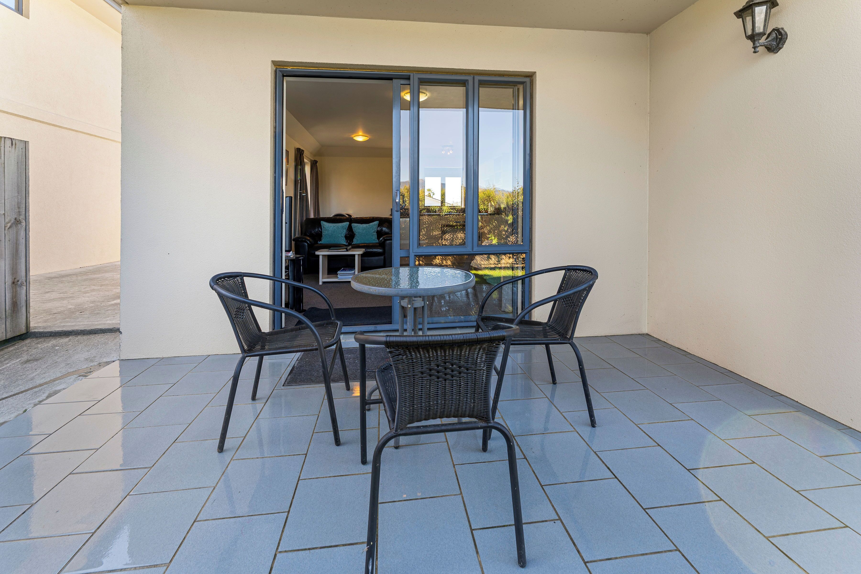 Une terrasse extérieure avec une table ronde et quatre chaises, une porte-fenêtre coulissante donnant sur un espace intérieur et un luminaire fixé au mur.