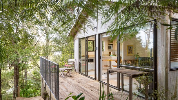A wooden deck with a glass-enclosed kitchen and dining area, surrounded by dense foliage.
