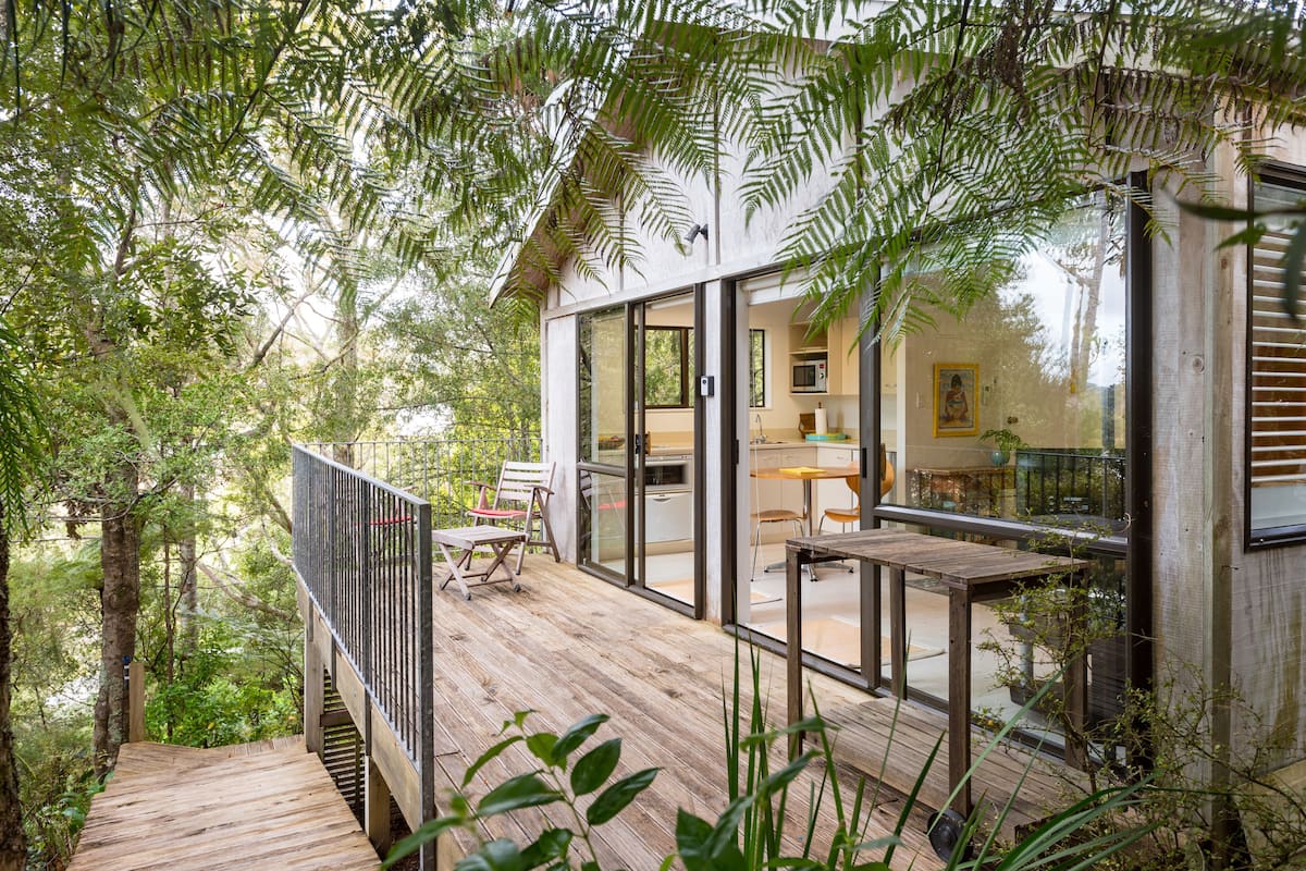 A wooden deck with a glass-enclosed kitchen and dining area, surrounded by dense foliage.