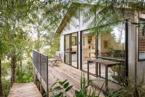 A wooden deck with a glass-enclosed kitchen and dining area, surrounded by dense foliage. - Craicor Accommodation (Paihia)