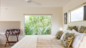 A bedroom with a large bed, a ceiling fan, a window with a view of greenery, and a small table with a floral bag.