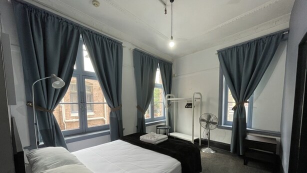 A hotel room with a bed, a bedside table, a desk, a chair, a standing fan, and a window with curtains. - The George Sydney - Hostel (Haymarket)