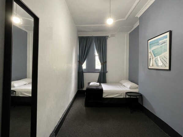 A hotel room with two single beds, a small table, a chair, a large mirror, a window with curtains, and a framed picture on the wall.