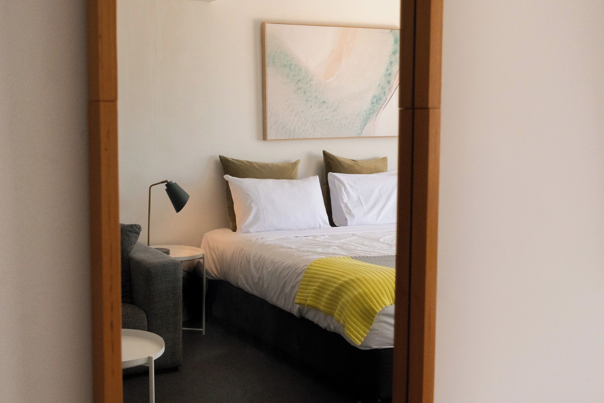 A hotel room with a bed, a bedside table, a lamp, a small chair, and a painting on the wall.