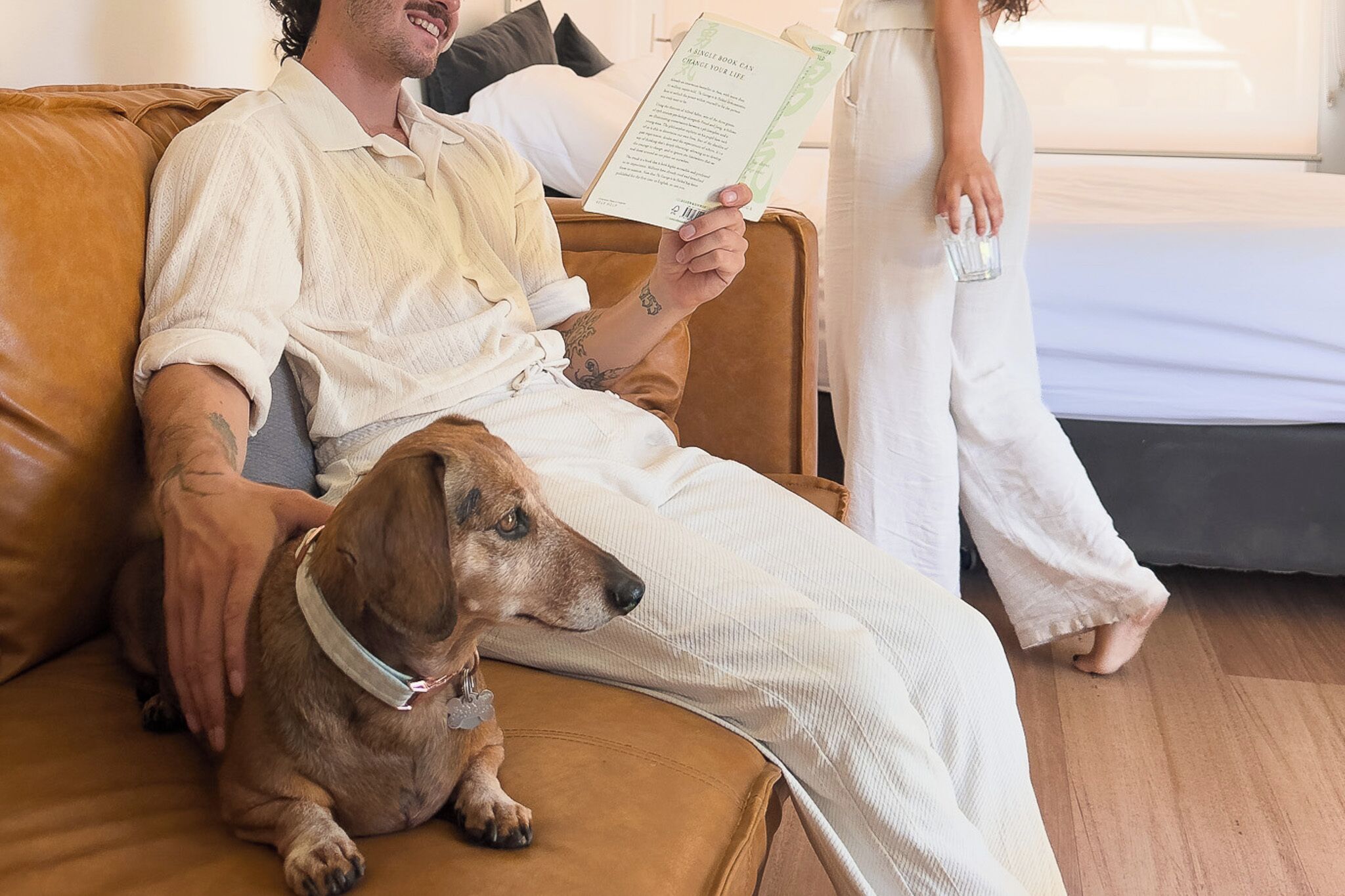 A man sitting on a brown leather couch with a dog, reading a book, and a woman standing beside him holding a glass.