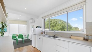 A modern kitchen with white cabinets, a large window, and a dining area with green chairs. - Moody's Motel (Blairgowrie)
