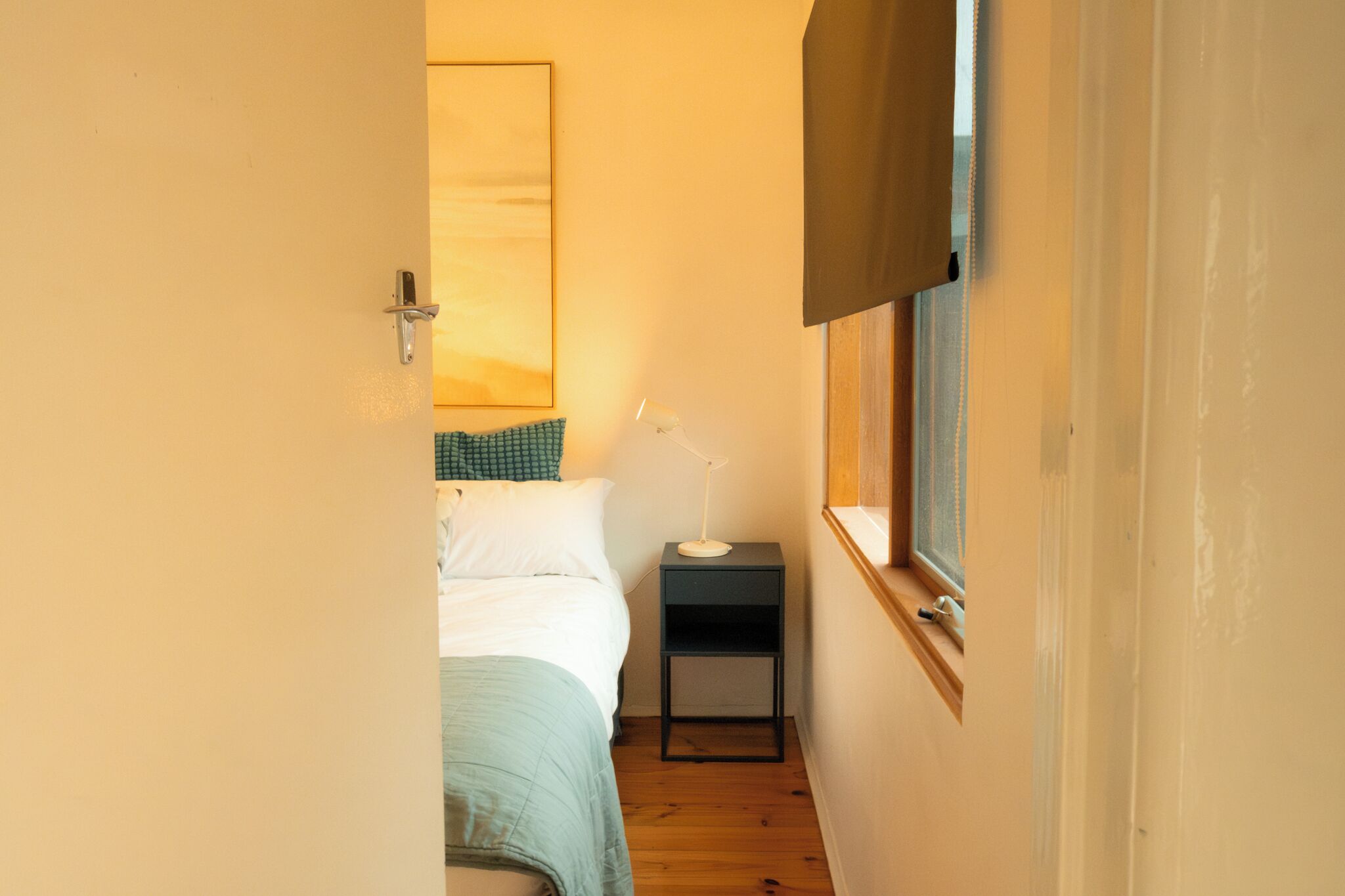 A narrow hallway leading to a bedroom with a bed, a nightstand, a lamp, and a window.