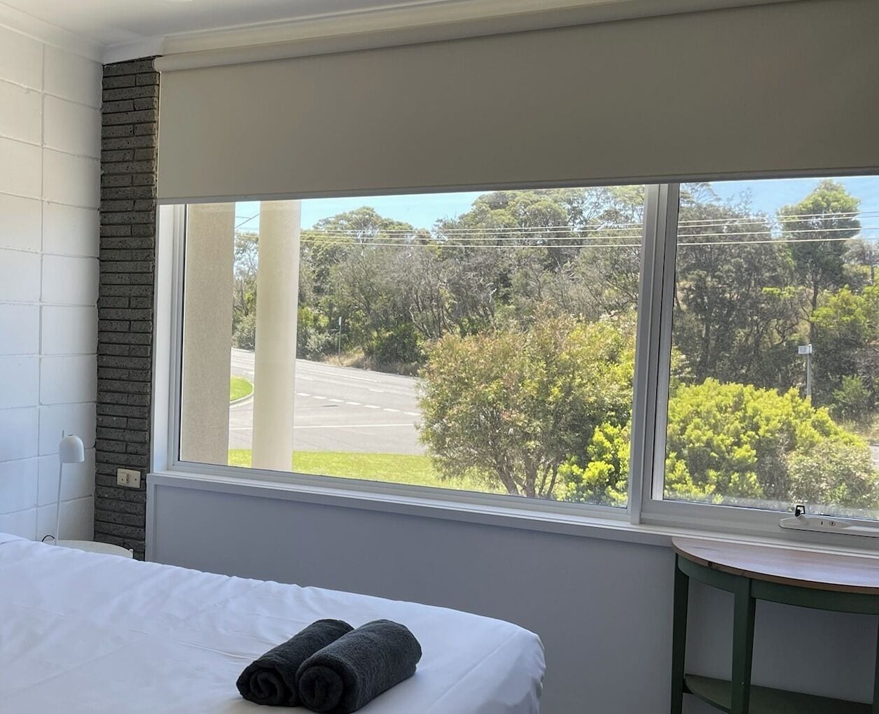 A hotel room with a bed, a window with a view of trees and a road, and a small table.