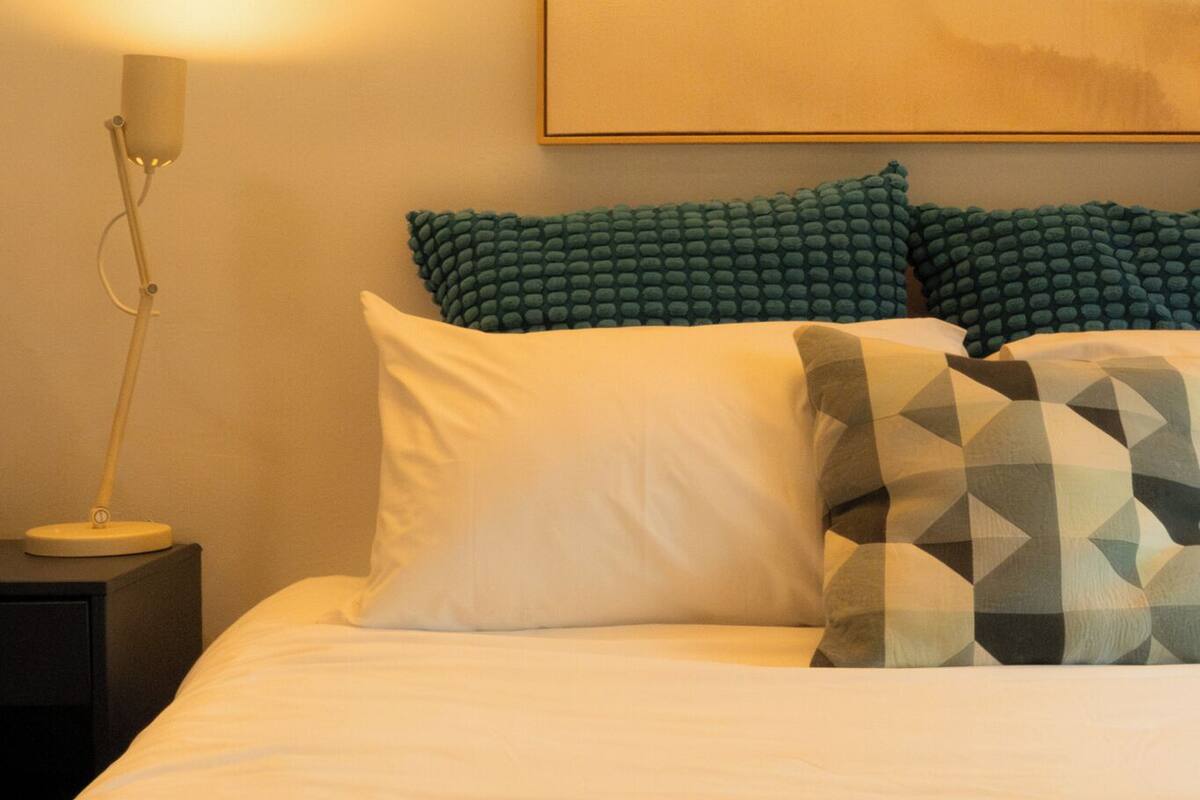 Uma cama bem arrumada com travesseiros, uma luminária de cabeceira e um livro aberto sobre a cama.