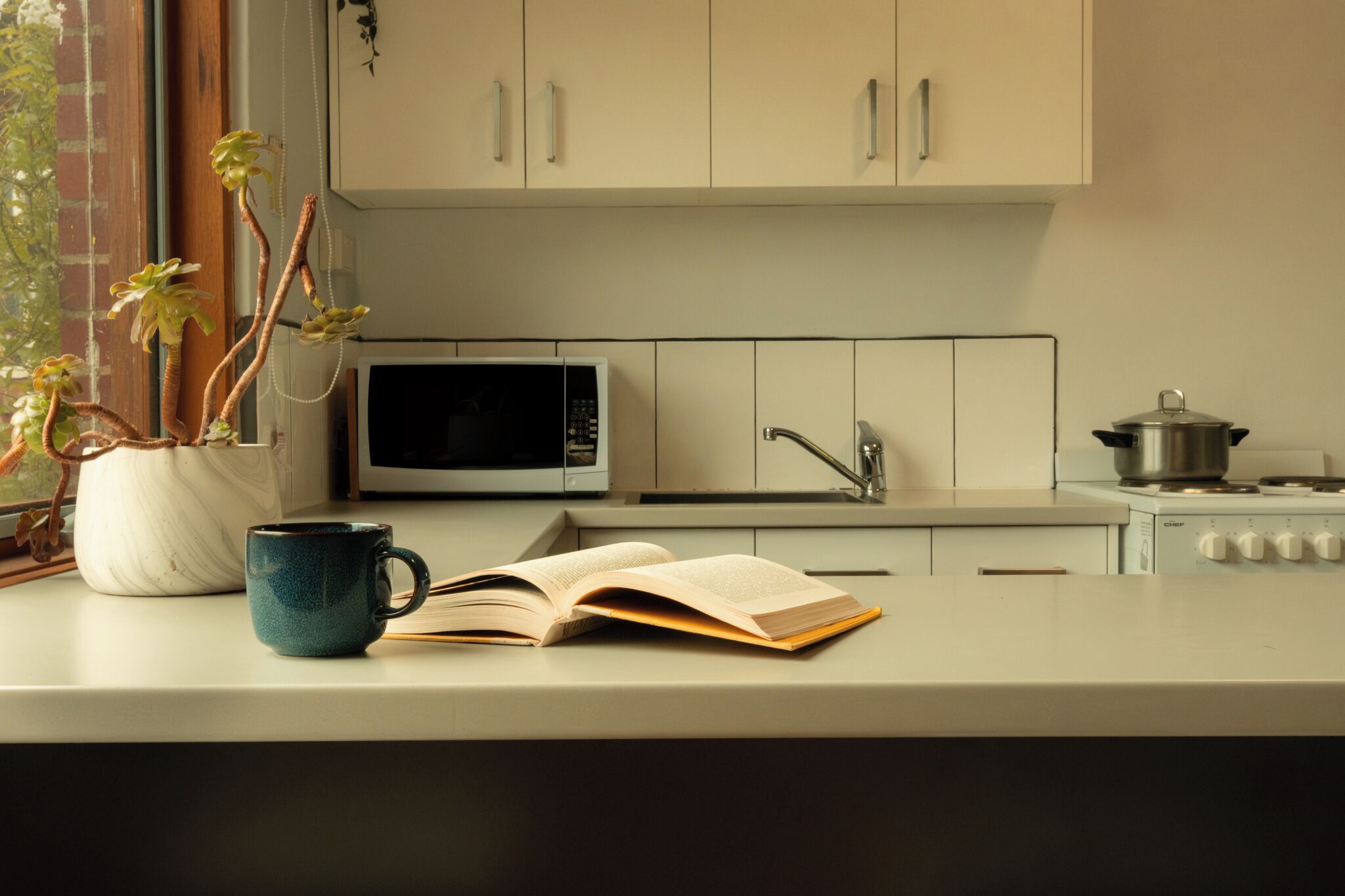 A kitchen with cabinets, a microwave, a stove, a coffee cup, and an open book.