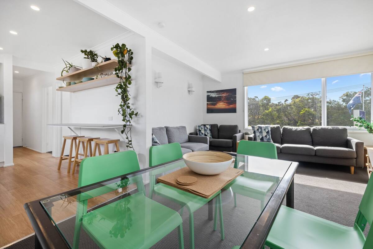Uma sala de estar moderna com uma mesa de jantar de vidro, cadeiras verdes e um sofá cinzento.