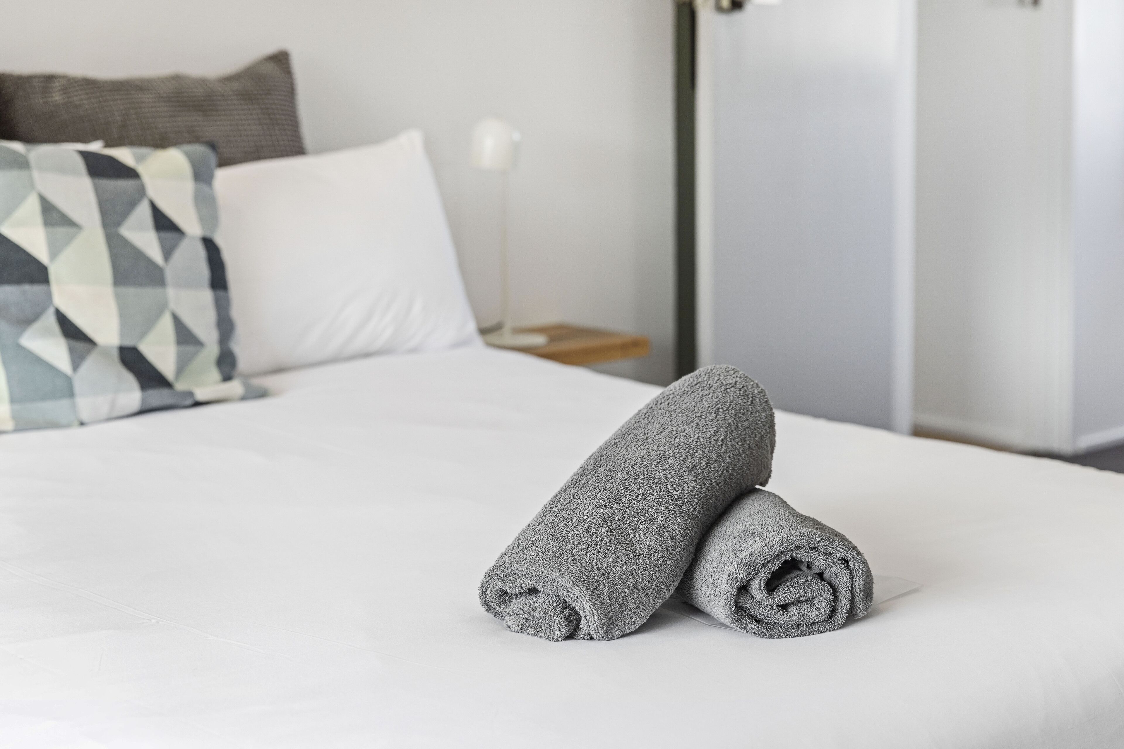 A neatly made bed with two rolled gray towels, a white pillow, and a geometric-patterned pillow on a white mattress.
