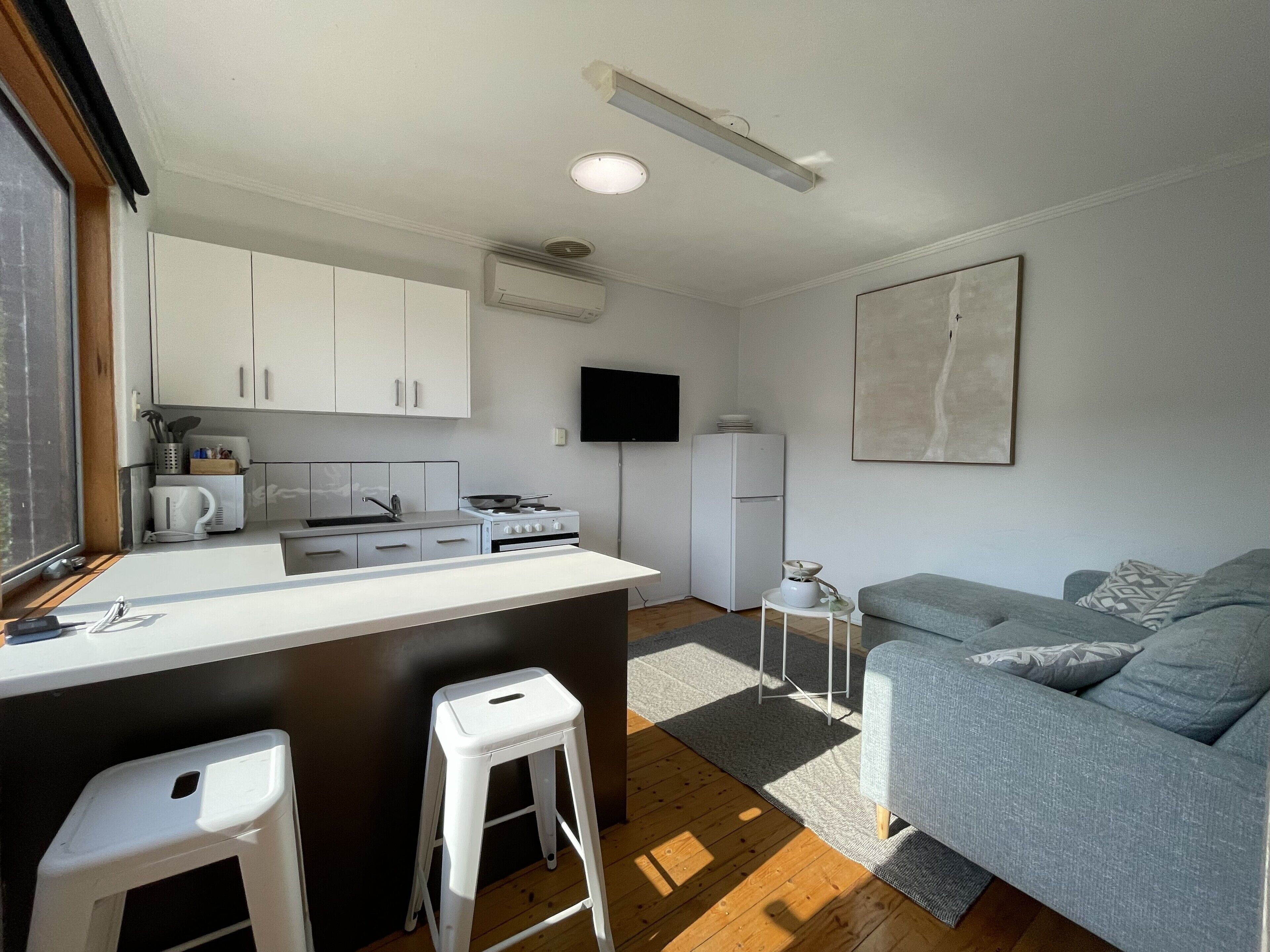A compact kitchen with white cabinets, a stove, and a sink. There's a small white stool and a gray couch in the living area.
