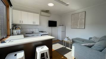 A compact kitchen with white cabinets, a stove, and a sink. There's a small white stool and a gray couch in the living area.