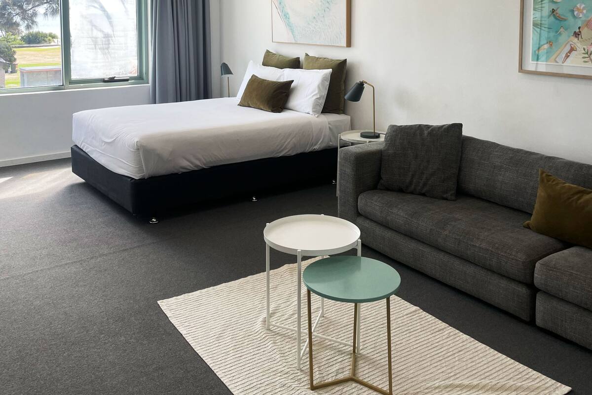 Quarto de hotel moderno com uma cama, um sofá cinzento, uma mesinha, uma janela grande e um aparelho de ar condicionado.