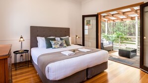 A bedroom with a large bed, a wooden nightstand, a small table with a lamp, and a view of a balcony with outdoor furniture. - Bay and Bush, Jervis Bay (Woollamia)