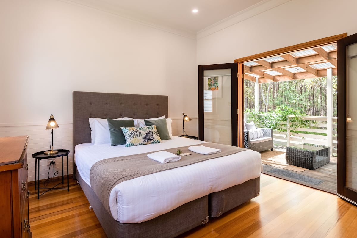A bedroom with a large bed, a wooden nightstand, a small table with a lamp, and a view of a balcony with outdoor furniture.