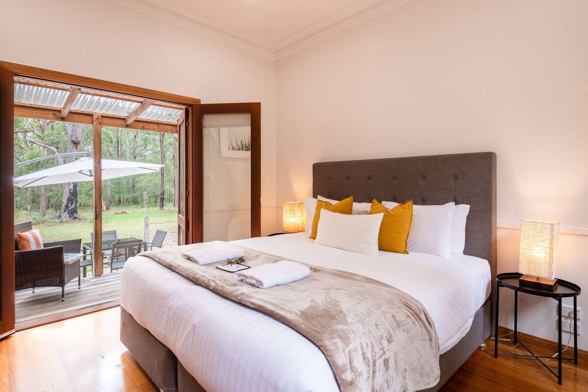 A large bed with a grey headboard, white bedding, and yellow pillows. There is a small table with a lamp beside the bed.
