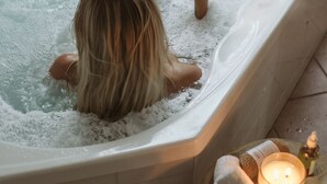 A person relaxing in a bathtub with bubbles, holding a bottle, and surrounded by a candle, a towel, and a small tray with toiletries. - Bay and Bush, Jervis Bay (Woollamia)