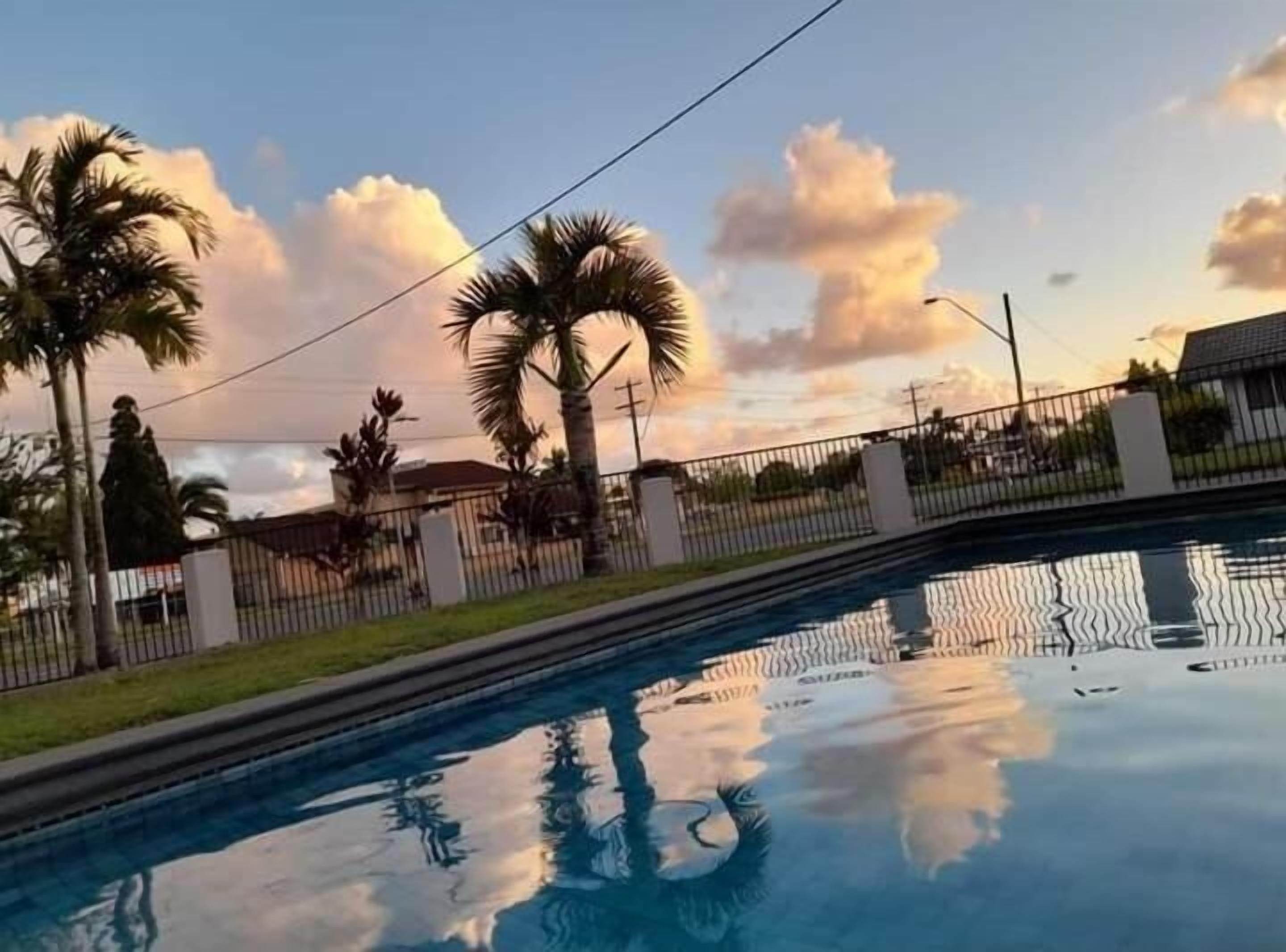 A swimming pool with a clear reflection, surrounded by a fence and palm trees, under a sunset sky.