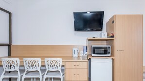A small kitchenette with a microwave, kettle, and a TV mounted on the wall. There are three white chairs and a wooden cabinet. - Casa Nostra Motel Mackay (West Mackay)