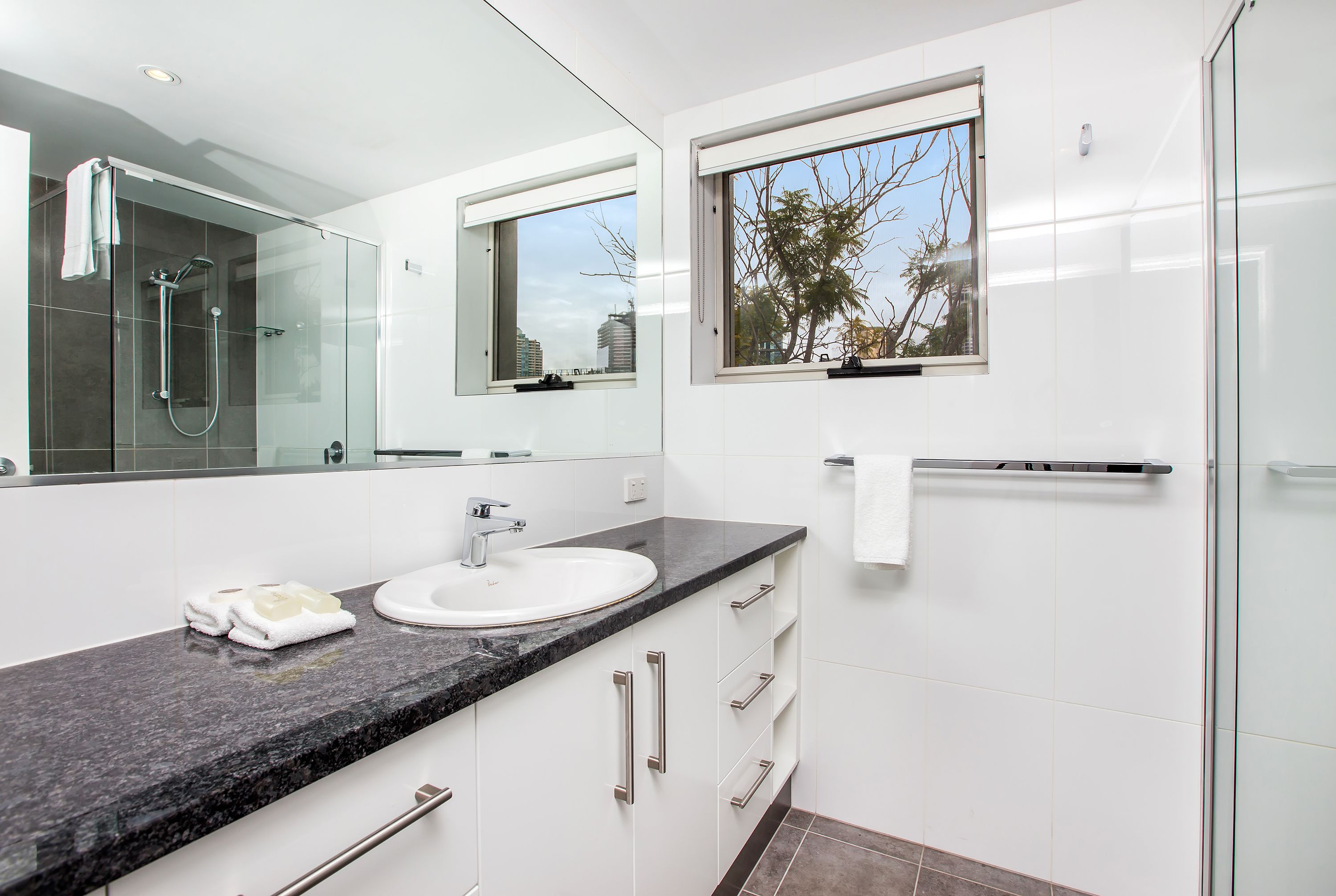 A modern bathroom with a glass shower enclosure, a large mirror, a sink with a marble countertop, and a window with a view of trees and buildings.
