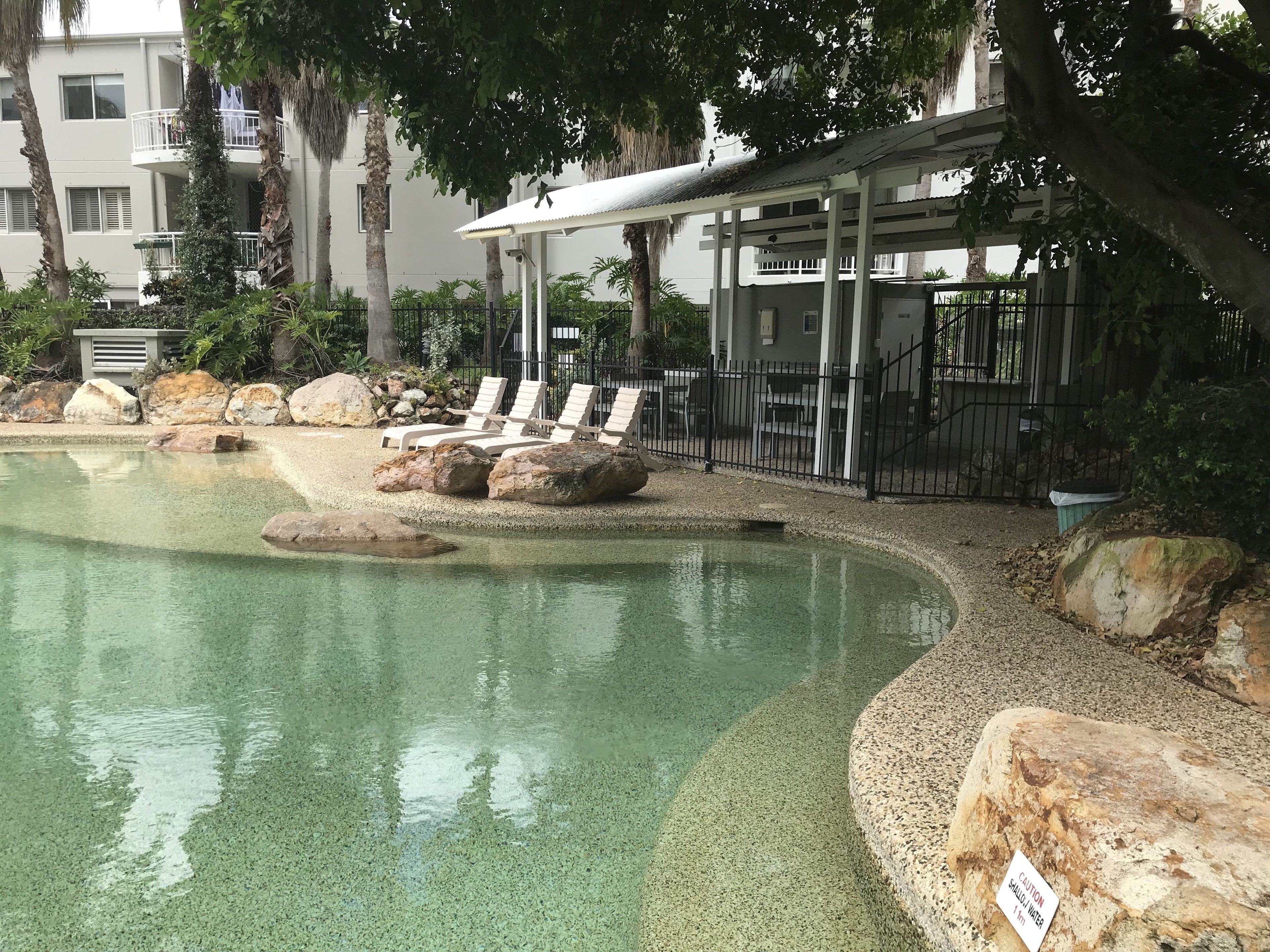 A swimming pool with lounge chairs and large rocks, surrounded by a fence and trees.