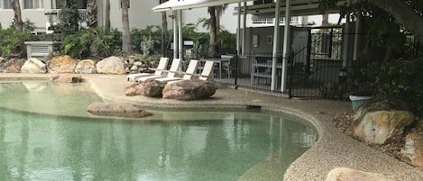En swimmingpool med solstolar och stora stenar, omgiven av ett staket och träd.