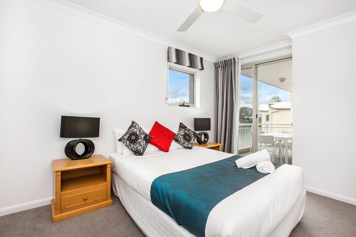 A modern hotel room with a large bed, a flat-screen TV, a wooden nightstand, and a balcony with a view of the city.