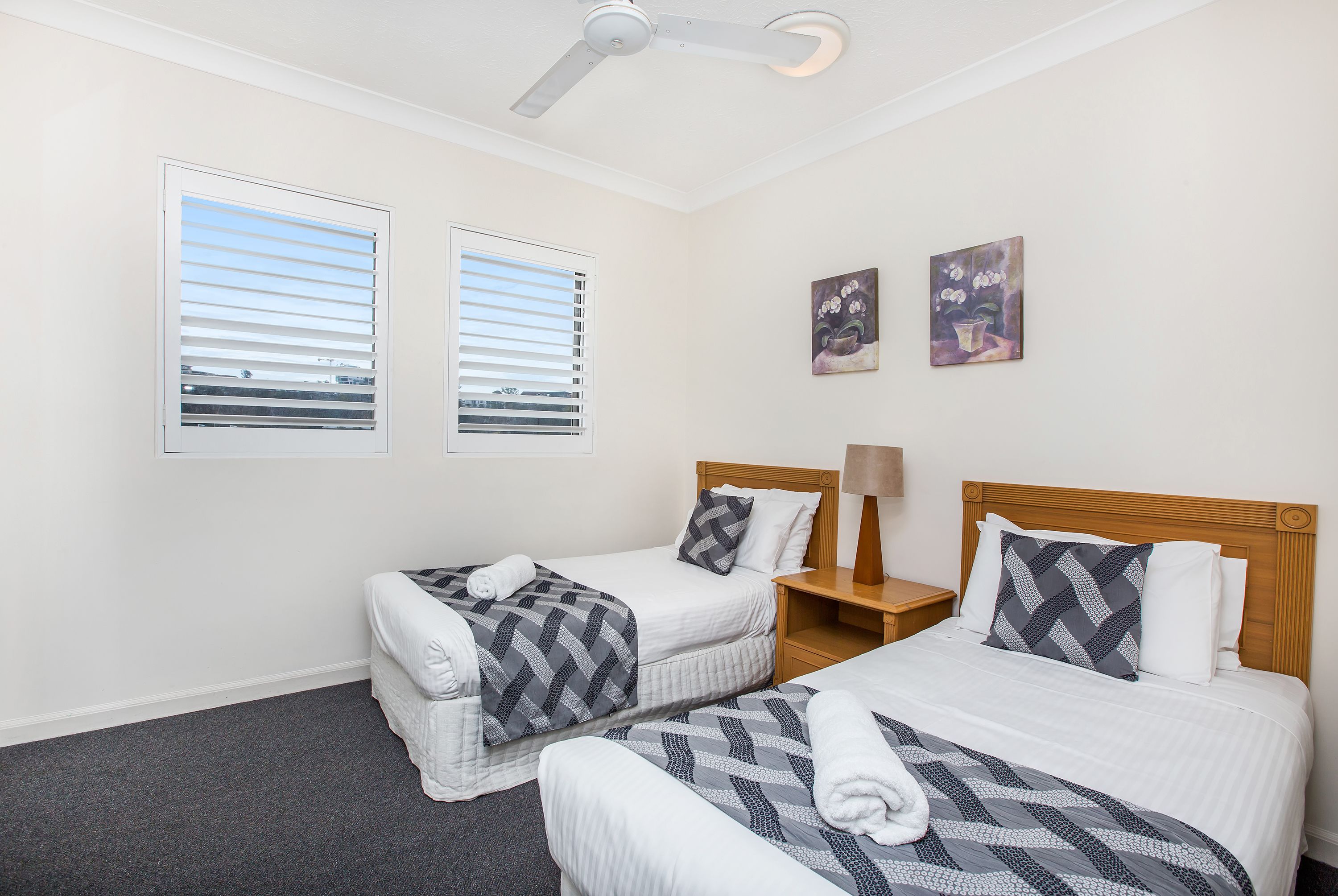 A hotel room with two single beds, a ceiling fan, two windows with blinds, a nightstand, and two wall-mounted paintings.
