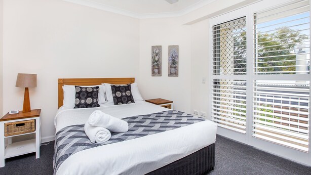A neatly made bed with a patterned blanket, a bedside lamp, and a small wicker basket on a nightstand. - Bridgewater Terraces (Kangaroo Point)