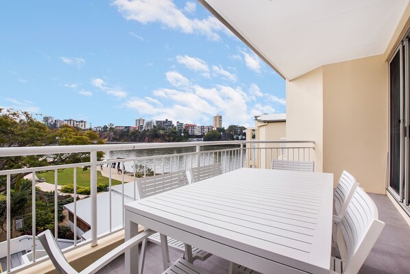 A balcony with a white table and chairs, overlooking a body of water and a cityscape.