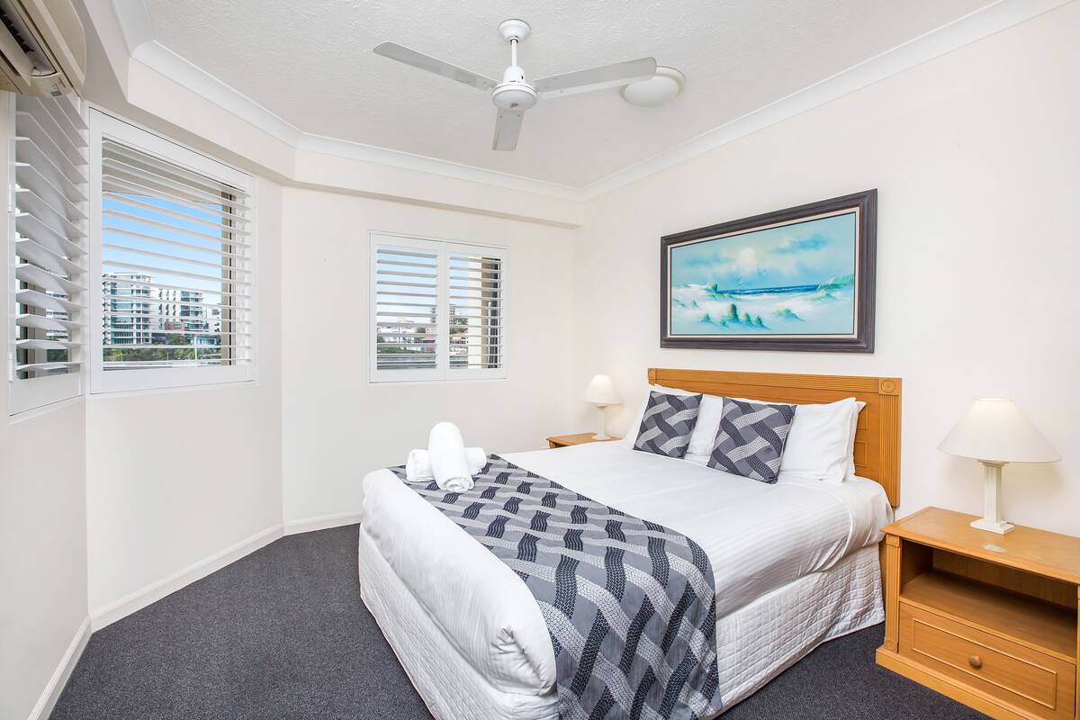A hotel room with a bed, two bedside tables, a ceiling fan, and a window with blinds.