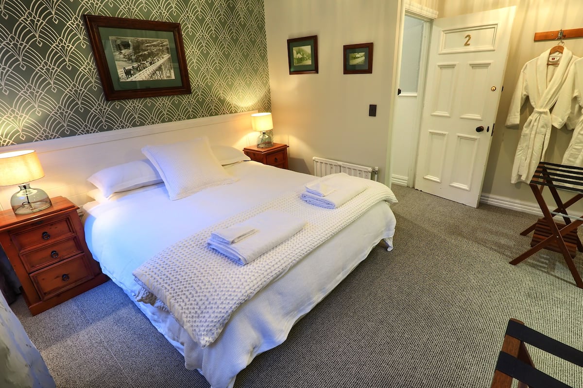 A neatly made bed with white linens and towels, a bedside table with a lamp, a wooden chair, and a door with two robes hanging on it.