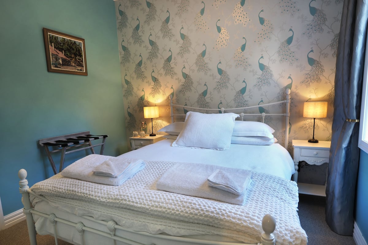 A neatly made bed with white linens and towels, a nightstand with a lamp, and a wall with peacock-patterned wallpaper.