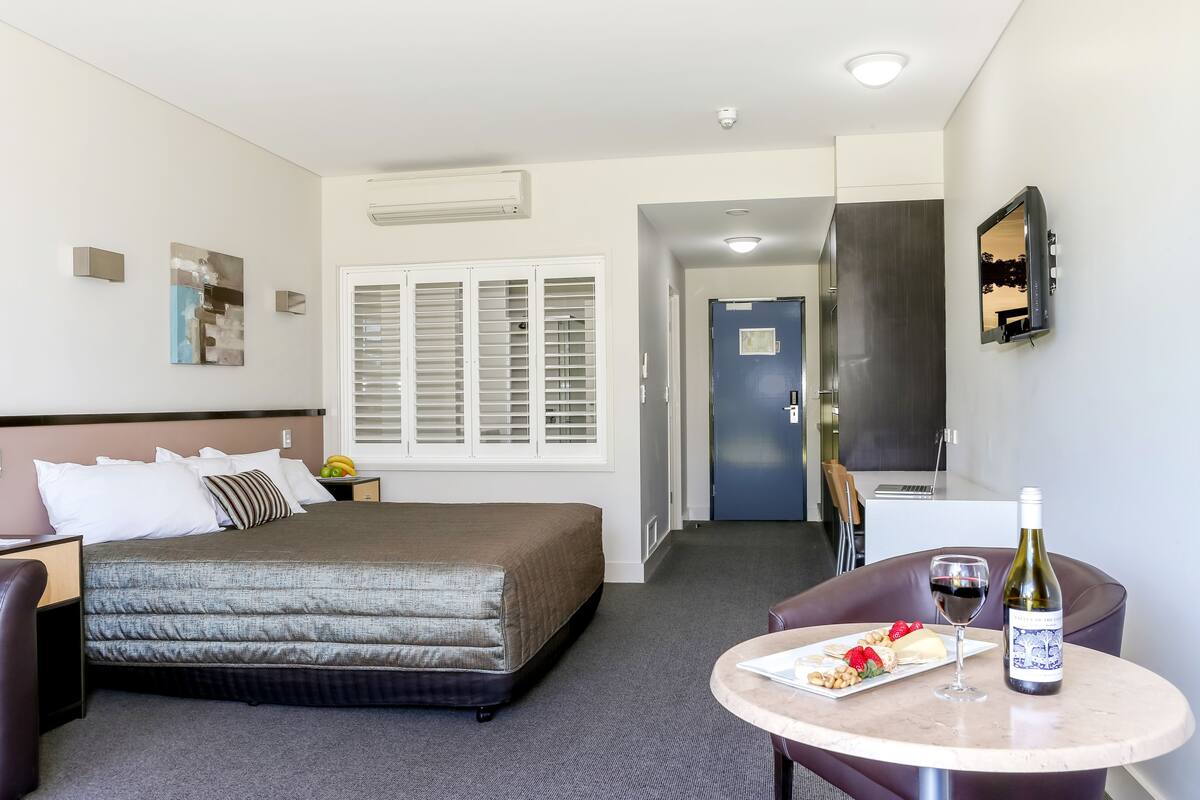 A hotel room with a bed, a dining table with a bottle of wine and a plate of food, a TV mounted on the wall, and a door leading to another room.