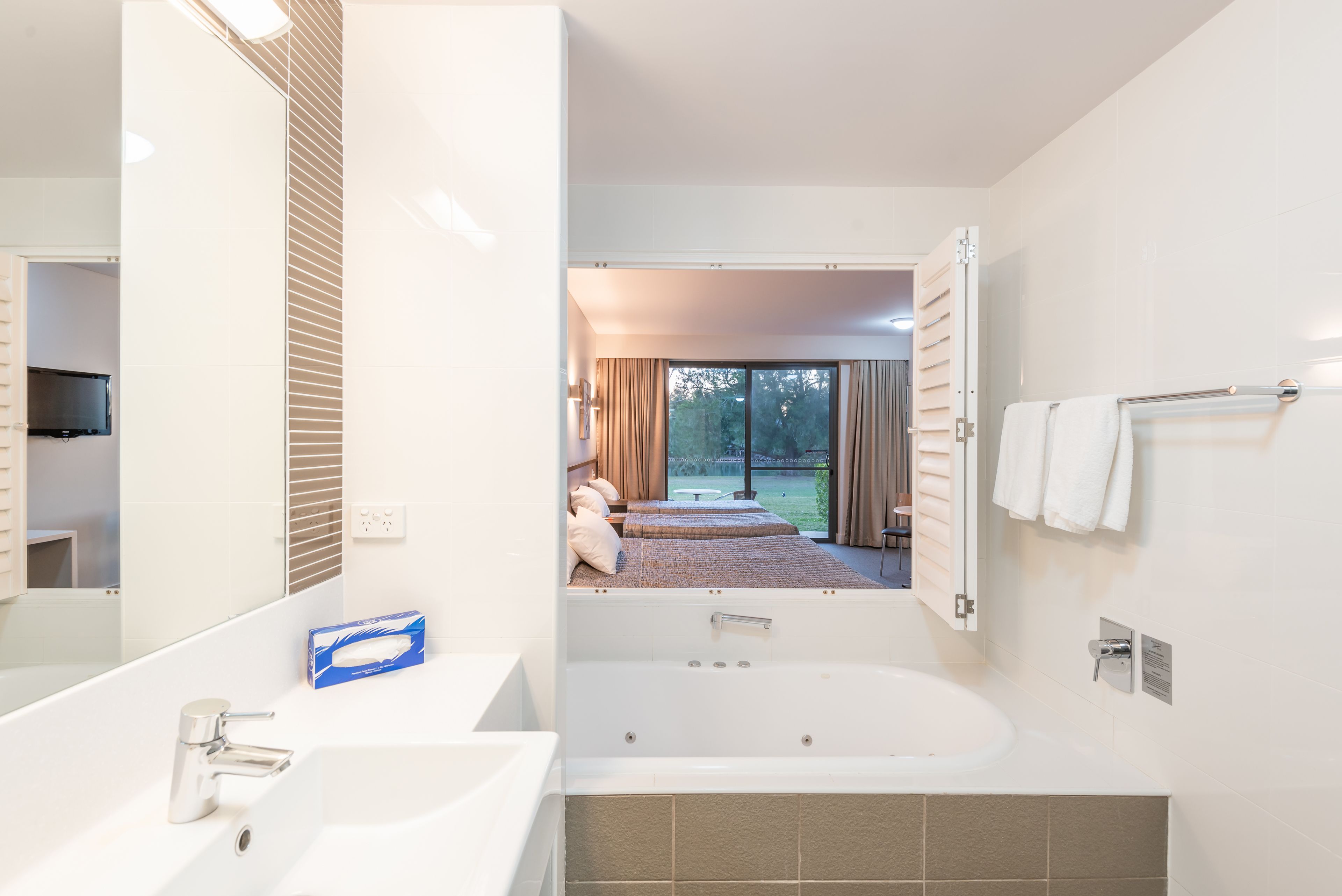 A modern bathroom with a large bathtub, a sink, and a mirror. There is a view of a bedroom with a bed and curtains.