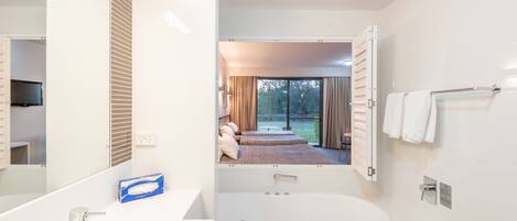 Un baño moderno con una bañera grande, un lavamanos y un espejo. Se ve una habitación con una cama y cortinas.