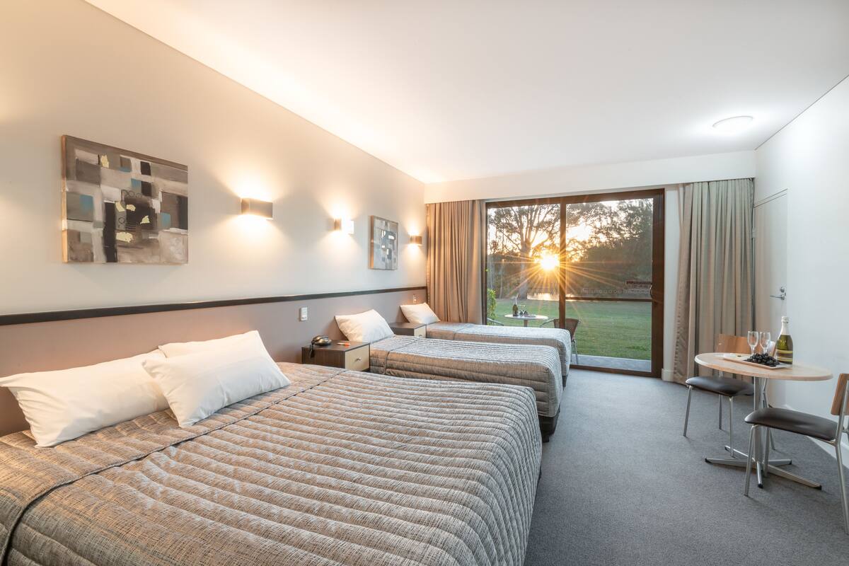 A hotel room with two beds, a small table with a bottle and glasses, and a view of the outdoors.
