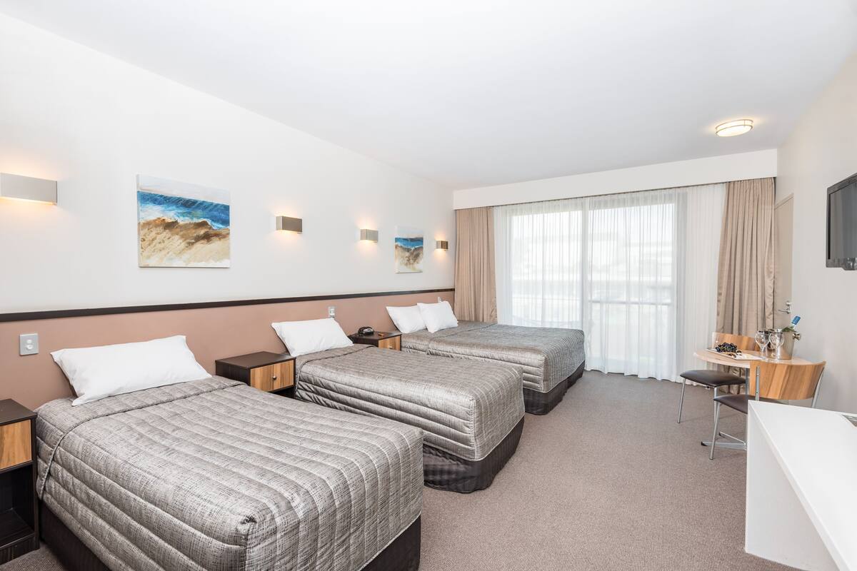 A hotel room with three beds, a desk, and a chair. There are wall-mounted lights and artwork.