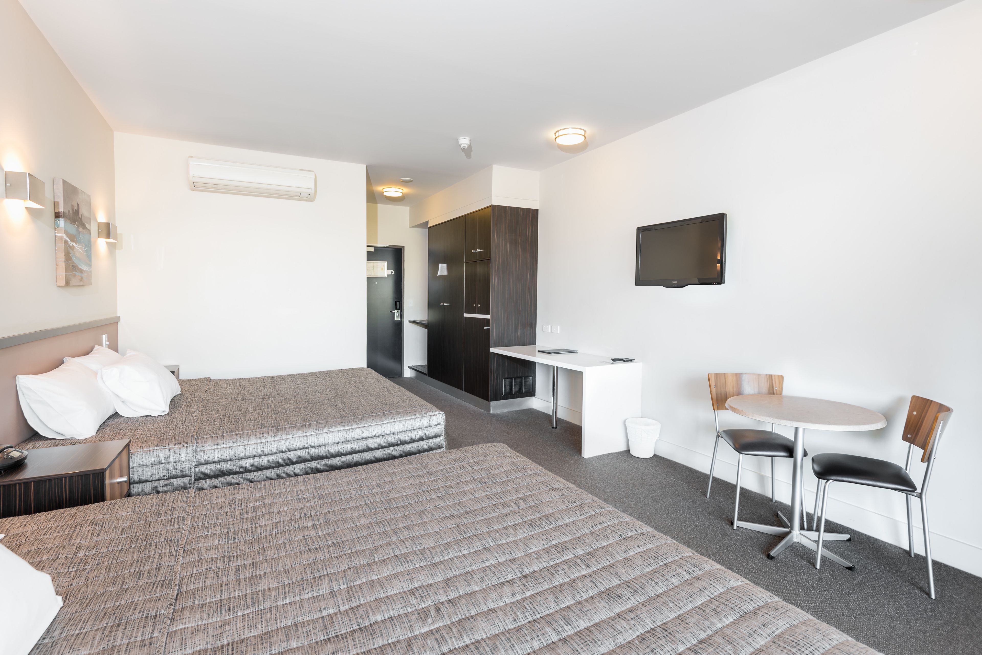 A hotel room with two beds, a desk, a chair, a TV, and a small kitchenette.