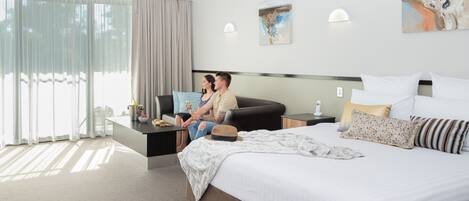 Una habitación de hotel moderna con una cama grande, un sofá, una mesa de comedor y un amplio ventanal con cortinas.