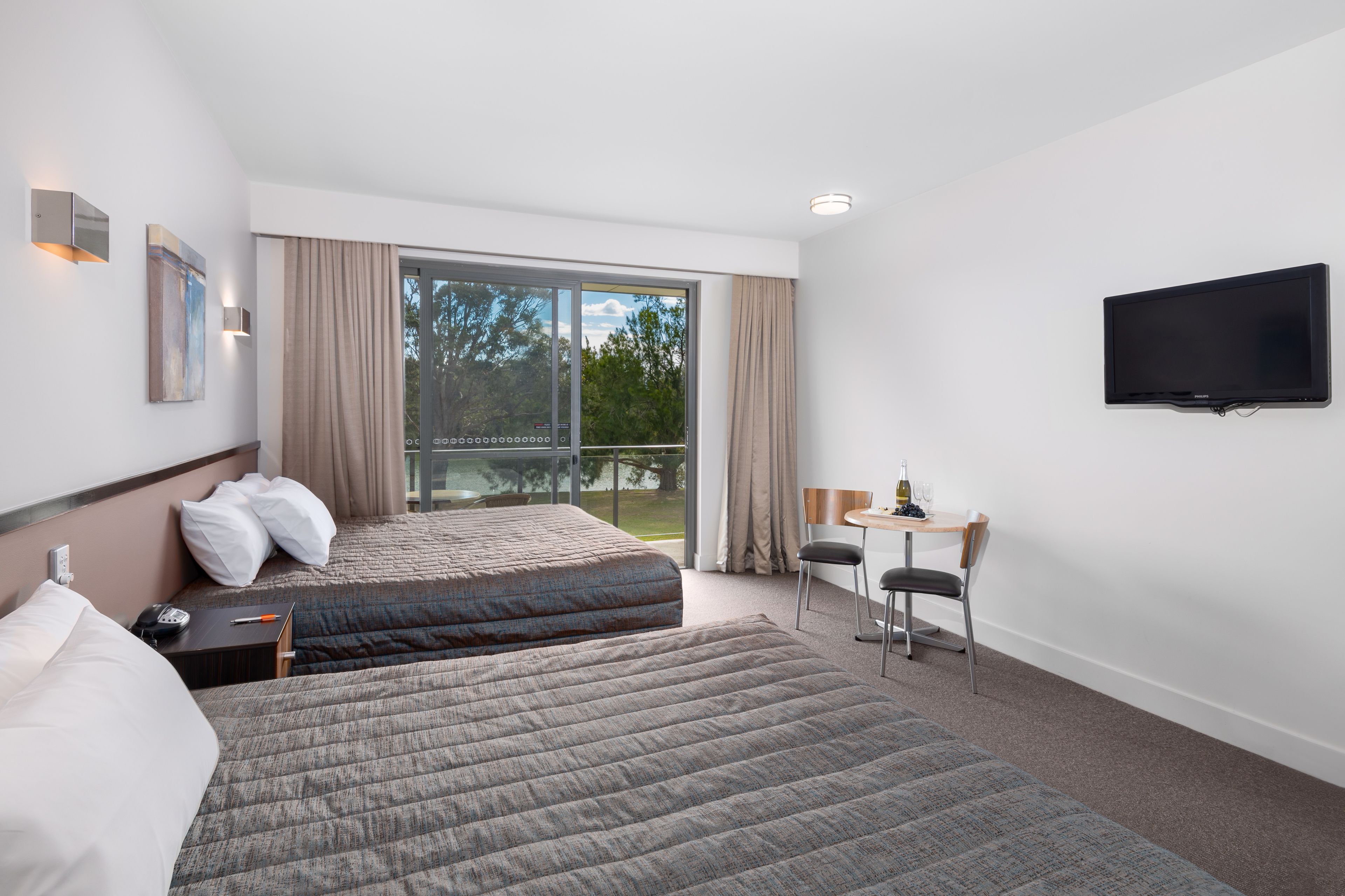 A hotel room with two beds, a desk with a chair, a flat-screen TV, and a large window with curtains.