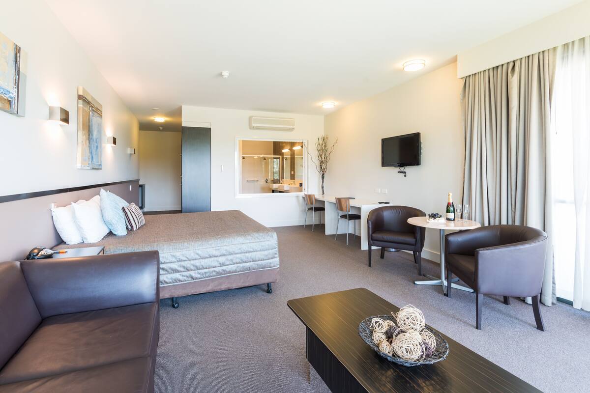 A hotel room with a bed, a sofa, a coffee table, a TV, and a dining area with chairs and a table.